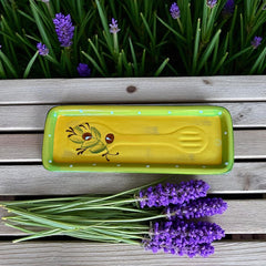 Rectangular ceramic spoon rest with yellow interior and green polka-dot rim, featuring hand-painted olive leaves, red berries, and whimsical fork and spoon motif; measures 11 by 4 inches.