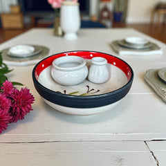 Round ceramic serving dish with red and black rim, hand-painted with green olive branches and brown olives on a cream base, accompanied by two small matching vessels nested inside.