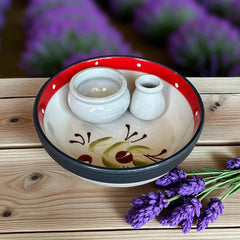 Round ceramic serving dish with red and black rim, hand-painted with green olive branches and brown olives on a cream base, accompanied by two small matching vessels nested inside.