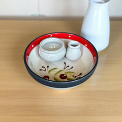 Round ceramic serving dish with red and black rim, hand-painted with green olive branches and brown olives on a cream base, accompanied by two small matching vessels nested inside.