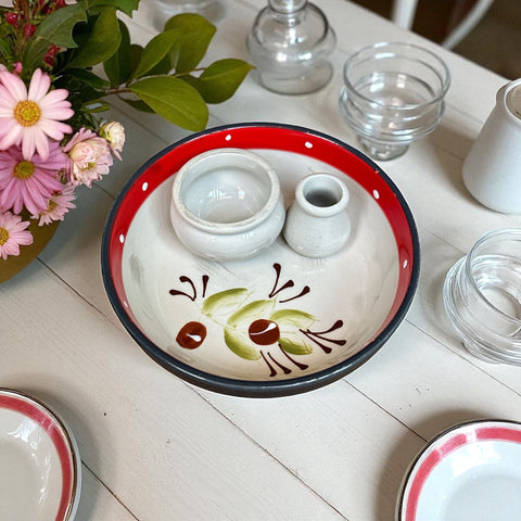 Round ceramic serving dish with red and black rim, hand-painted with green olive branches and brown olives on a cream base, accompanied by two small matching vessels nested inside.