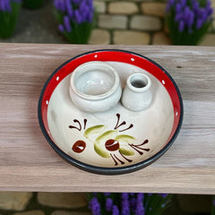 Round ceramic serving dish with red and black rim, hand-painted with green olive branches and brown olives on a cream base, accompanied by two small matching vessels nested inside.