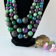 Vintage 1950s three‑strand necklace with green, purple, black, and blue graduated plastic beads and gold‑tone connectors displayed on neutral background.