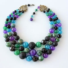 Vintage 1950s three‑strand necklace with green, purple, black, and blue graduated plastic beads and gold‑tone connectors displayed on neutral background.