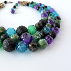 Vintage 1950s three‑strand necklace with green, purple, black, and blue graduated plastic beads and gold‑tone connectors displayed on neutral background.