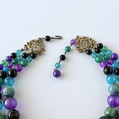 Vintage 1950s three‑strand necklace with green, purple, black, and blue graduated plastic beads and gold‑tone connectors displayed on neutral background.