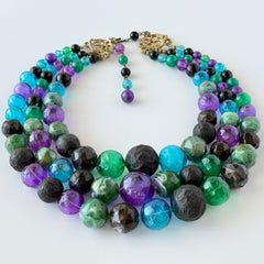 Vintage 1950s three‑strand necklace with green, purple, black, and blue graduated plastic beads and gold‑tone connectors displayed on neutral background.