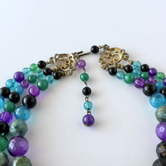 Vintage 1950s three‑strand necklace with green, purple, black, and blue graduated plastic beads and gold‑tone connectors displayed on neutral background.