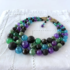 Vintage 1950s three‑strand necklace with green, purple, black, and blue graduated plastic beads and gold‑tone connectors displayed on neutral background.