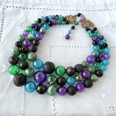 Vintage 1950s three‑strand necklace with green, purple, black, and blue graduated plastic beads and gold‑tone connectors displayed on neutral background.