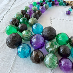Vintage 1950s three‑strand necklace with green, purple, black, and blue graduated plastic beads and gold‑tone connectors displayed on neutral background.
