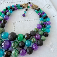 Vintage 1950s three‑strand necklace with green, purple, black, and blue graduated plastic beads and gold‑tone connectors displayed on neutral background.