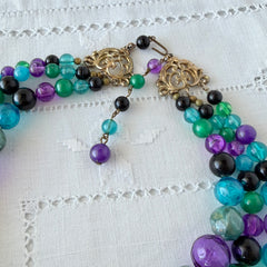Vintage 1950s three‑strand necklace with green, purple, black, and blue graduated plastic beads and gold‑tone connectors displayed on neutral background.