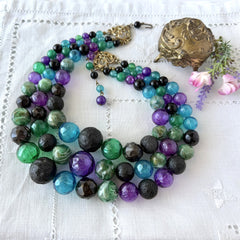 Vintage 1950s three‑strand necklace with green, purple, black, and blue graduated plastic beads and gold‑tone connectors displayed on neutral background.