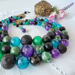 Vintage 1950s three‑strand necklace with green, purple, black, and blue graduated plastic beads and gold‑tone connectors displayed on neutral background.