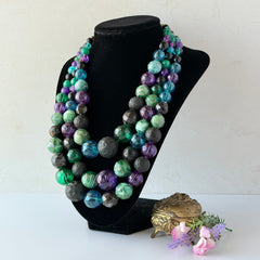Vintage 1950s three‑strand necklace with green, purple, black, and blue graduated plastic beads and gold‑tone connectors displayed on neutral background.