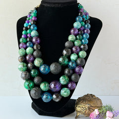 Vintage 1950s three‑strand necklace with green, purple, black, and blue graduated plastic beads and gold‑tone connectors displayed on neutral background.