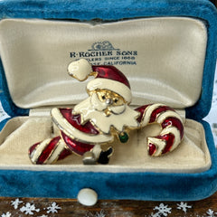 Vintage gold-tone brooch featuring Santa Claus in red and white enamel, riding a striped candy cane with green accent; 1980s/90s holiday design, photographed on neutral background.