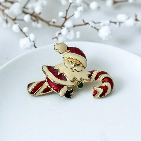 Vintage gold-tone brooch featuring Santa Claus in red and white enamel, riding a striped candy cane with green accent; 1980s/90s holiday design, photographed on neutral background.