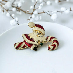 Vintage gold-tone brooch featuring Santa Claus in red and white enamel, riding a striped candy cane with green accent; 1980s/90s holiday design, photographed on neutral background.