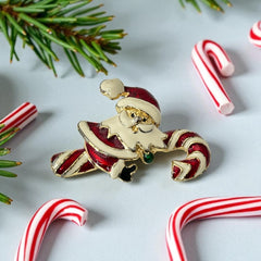Vintage gold-tone brooch featuring Santa Claus in red and white enamel, riding a striped candy cane with green accent; 1980s/90s holiday design, photographed on neutral background.