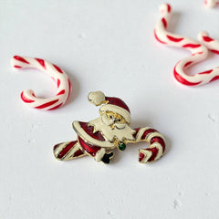 Vintage gold-tone brooch featuring Santa Claus in red and white enamel, riding a striped candy cane with green accent; 1980s/90s holiday design, photographed on neutral background.