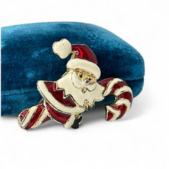 Vintage gold-tone brooch featuring Santa Claus in red and white enamel, riding a striped candy cane with green accent; 1980s/90s holiday design, photographed on neutral background.
