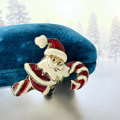Vintage gold-tone brooch featuring Santa Claus in red and white enamel, riding a striped candy cane with green accent; 1980s/90s holiday design, photographed on neutral background.