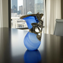 Signed Romanian handblown art glass vase from the 1990s featuring swirling blue glass and sculptural bronze overlay with organic vine-like forms. The vase has a flared top, narrow base, and an undeciphered artist’s signature etched into the glass. A collectible decorative piece in very good vintage condition.