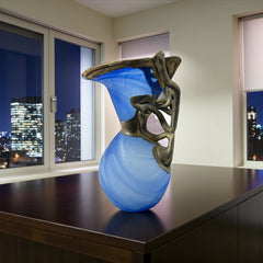 Signed Romanian handblown art glass vase from the 1990s featuring swirling blue glass and sculptural bronze overlay with organic vine-like forms. The vase has a flared top, narrow base, and an undeciphered artist’s signature etched into the glass. A collectible decorative piece in very good vintage condition.
