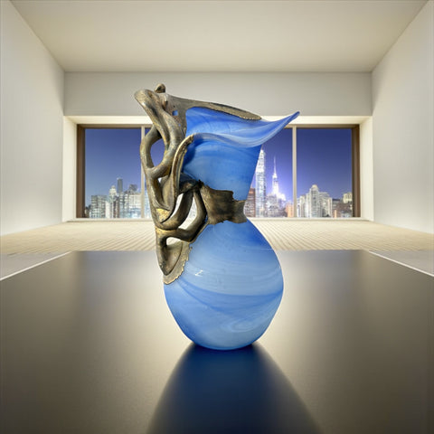 Signed Romanian handblown art glass vase from the 1990s featuring swirling blue glass and sculptural bronze overlay with organic vine-like forms. The vase has a flared top, narrow base, and an undeciphered artist’s signature etched into the glass. A collectible decorative piece in very good vintage condition.