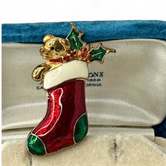 Vintage SFJ Christmas stocking brooch in gold tone metal with red, green, and white enamel; features teddy bear and holly details, signed SFJ on back.