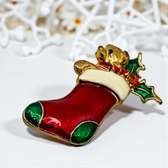 Vintage SFJ Christmas stocking brooch in gold tone metal with red, green, and white enamel; features teddy bear and holly details, signed SFJ on back.