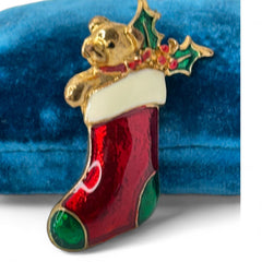 Vintage SFJ Christmas stocking brooch in gold tone metal with red, green, and white enamel; features teddy bear and holly details, signed SFJ on back.