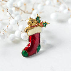 Vintage SFJ Christmas stocking brooch in gold tone metal with red, green, and white enamel; features teddy bear and holly details, signed SFJ on back.