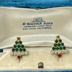 Pair of vintage Sarah Coventry clip-on earrings shaped like Christmas trees, featuring clear and green rhinestones as leaves, a red rhinestone trunk, and a yellow rhinestone star at the top. Gold tone metal with signed clips. Small size, festive design.