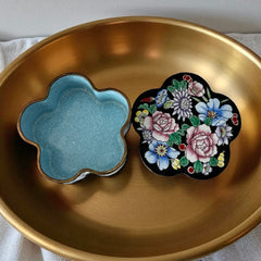 Vintage Seymour Mann flower-shaped trinket box with black enamel exterior, decorated with multicolored floral designs outlined in gold; features a light blue enamel interior and sculpted petal edges.
