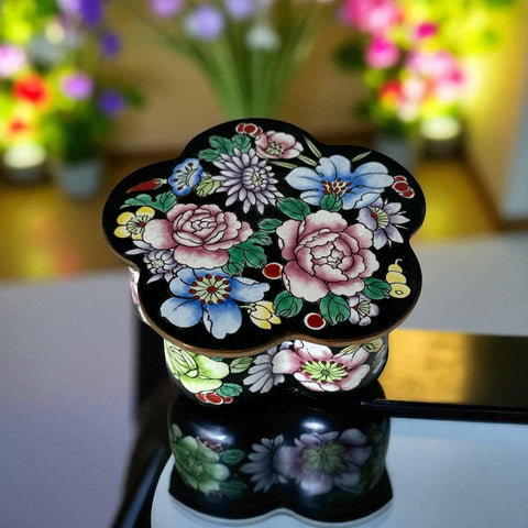 Vintage Seymour Mann flower-shaped trinket box with black enamel exterior, decorated with multicolored floral designs outlined in gold; features a light blue enamel interior and sculpted petal edges.