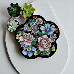 Vintage Seymour Mann flower-shaped trinket box with black enamel exterior, decorated with multicolored floral designs outlined in gold; features a light blue enamel interior and sculpted petal edges.
