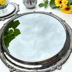 Vintage Sheffield Silver Co. Shrewsbury Plateau round silver-plated tray with mirrored surface and ornate floral scrollwork border; footed design with four decorative legs; shown outdoors with sky and leaves reflected in mirror; very good vintage condition with original packaging and maker’s sticker from Norton, Massachusetts.