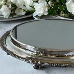 Vintage Sheffield Silver Co. Shrewsbury Plateau round silver-plated tray with mirrored surface and ornate floral scrollwork border; footed design with four decorative legs; shown outdoors with sky and leaves reflected in mirror; very good vintage condition with original packaging and maker’s sticker from Norton, Massachusetts.