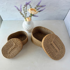Two oval sweetgrass baskets with lids, hand‑woven in natural tones, shown nested together beside a white vase with flowers.