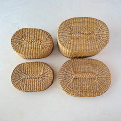 Two oval sweetgrass baskets with lids, hand‑woven in natural tones, shown nested together beside a white vase with flowers.