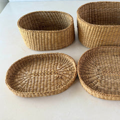 Two oval sweetgrass baskets with lids, hand‑woven in natural tones, shown nested together beside a white vase with flowers.