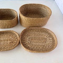Two oval sweetgrass baskets with lids, hand‑woven in natural tones, shown nested together beside a white vase with flowers.