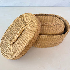 Two oval sweetgrass baskets with lids, hand‑woven in natural tones, shown nested together beside a white vase with flowers.