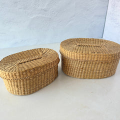 Two oval sweetgrass baskets with lids, hand‑woven in natural tones, shown nested together beside a white vase with flowers.