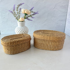 Two oval sweetgrass baskets with lids, hand‑woven in natural tones, shown nested together beside a white vase with flowers.