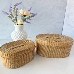 Two oval sweetgrass baskets with lids, hand‑woven in natural tones, shown nested together beside a white vase with flowers.