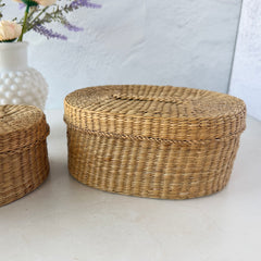 Two oval sweetgrass baskets with lids, hand‑woven in natural tones, shown nested together beside a white vase with flowers.